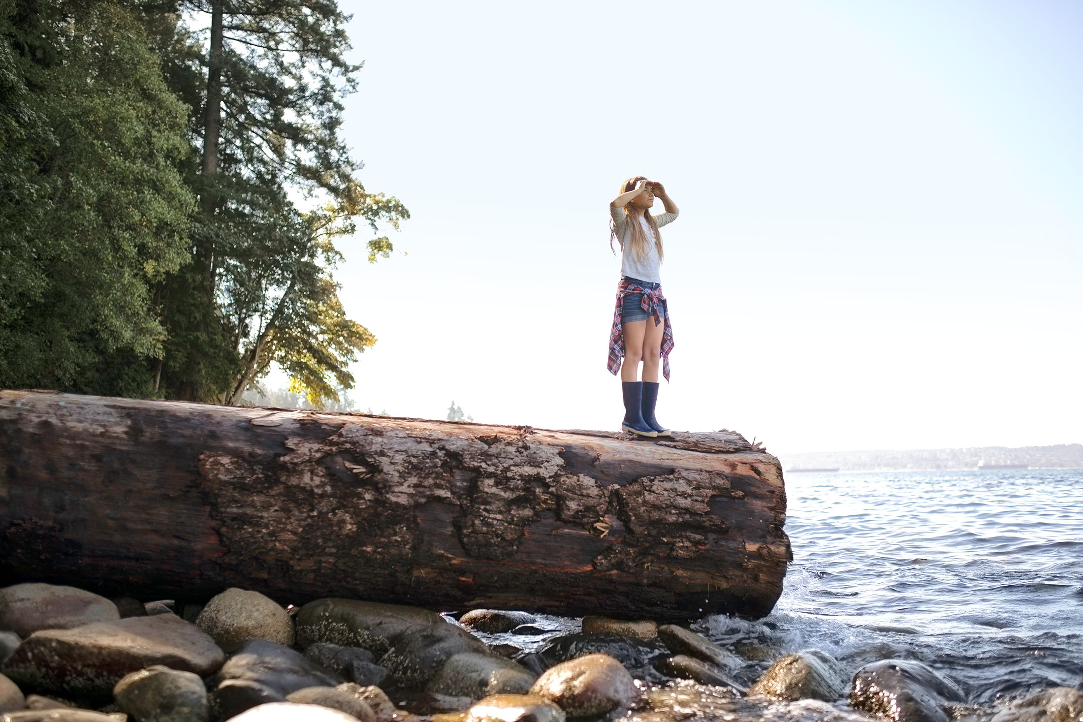 Mädchen steht auf einem Baumstamm am Meer und schaut aufs Wasser