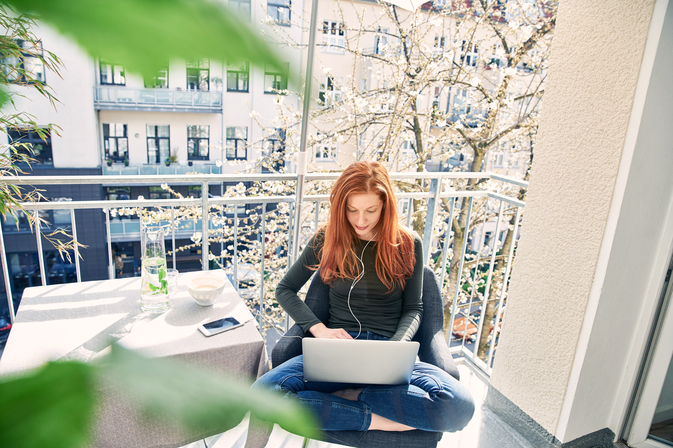 Rothaarige junge Frau sitzt auf dem Balkon und arbeitet am Laptop
