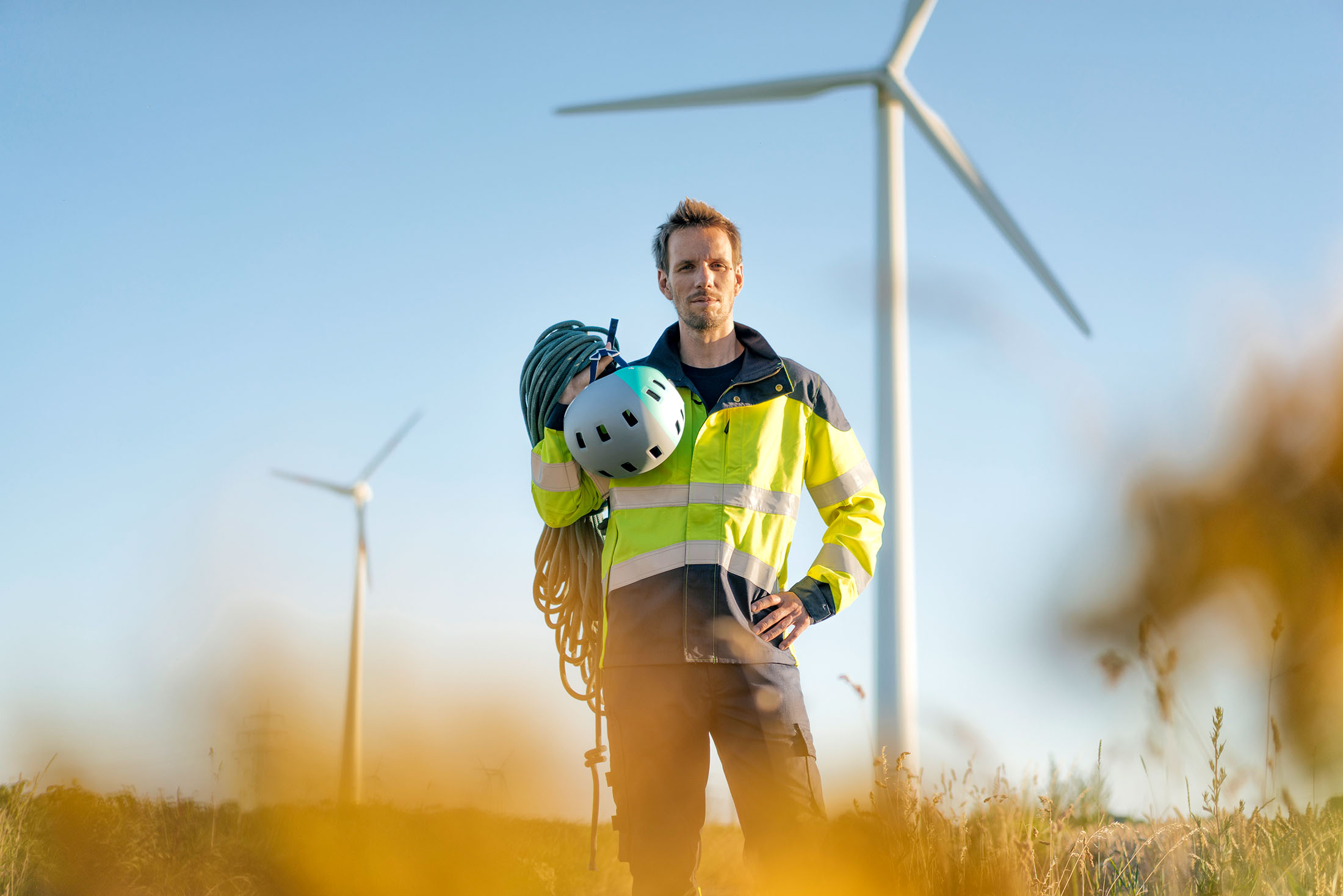 Techniker steht mit Kletterausrüstung auf einem Feld, im Hintergund Windräder