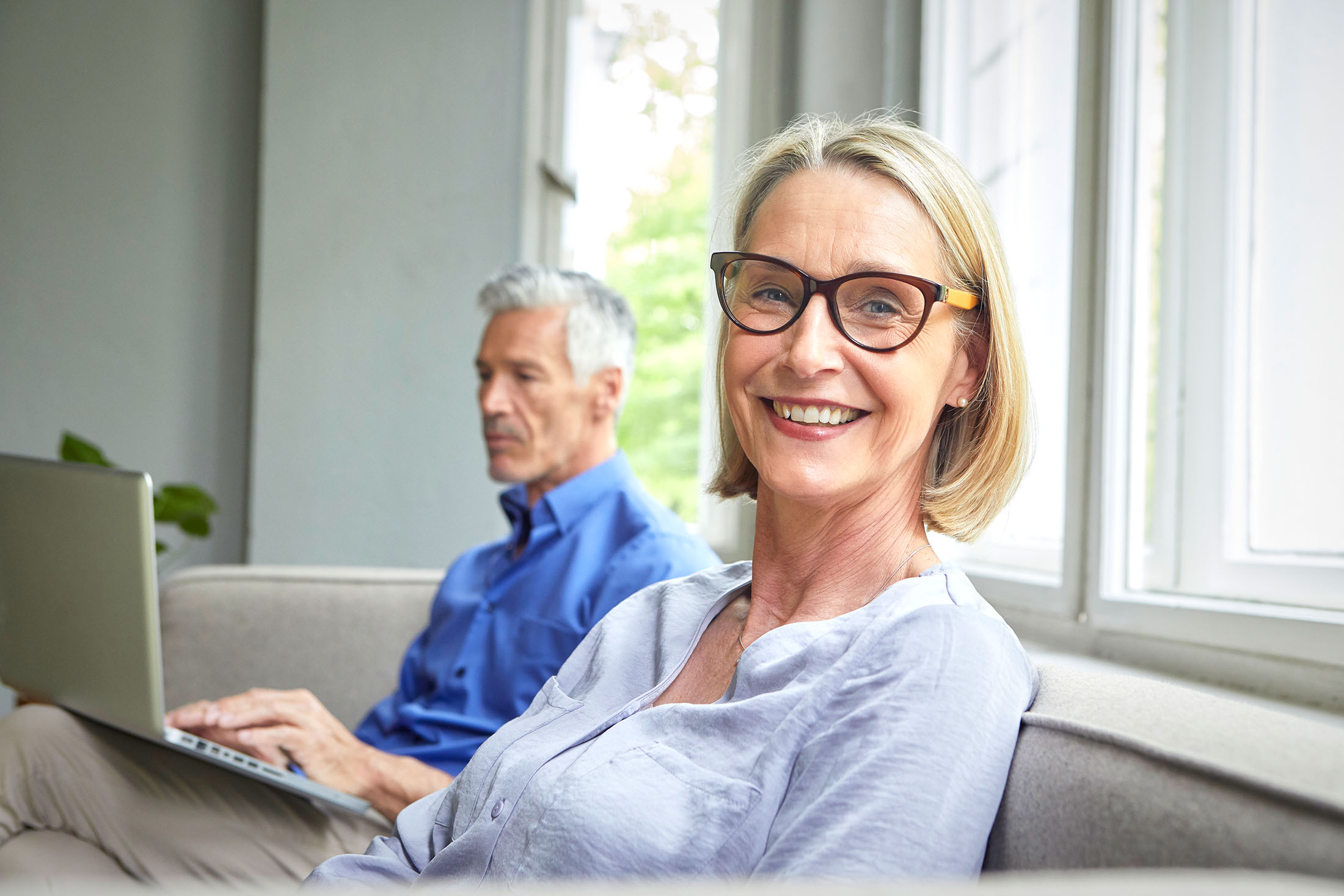 Portait einer älteren lächelnden Frau mit Mann im Hintergrund am Laptop der ein Laptop benutzt