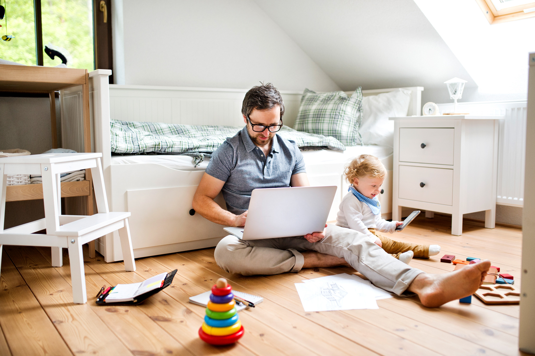Ein Vater sitzt vor einem Bett auf dem Boden und arbeitet mit dem Laptop. Sein Kleinkind sitzt neben ihm und schaut lächelnd auf Spielzeig vor ihm.