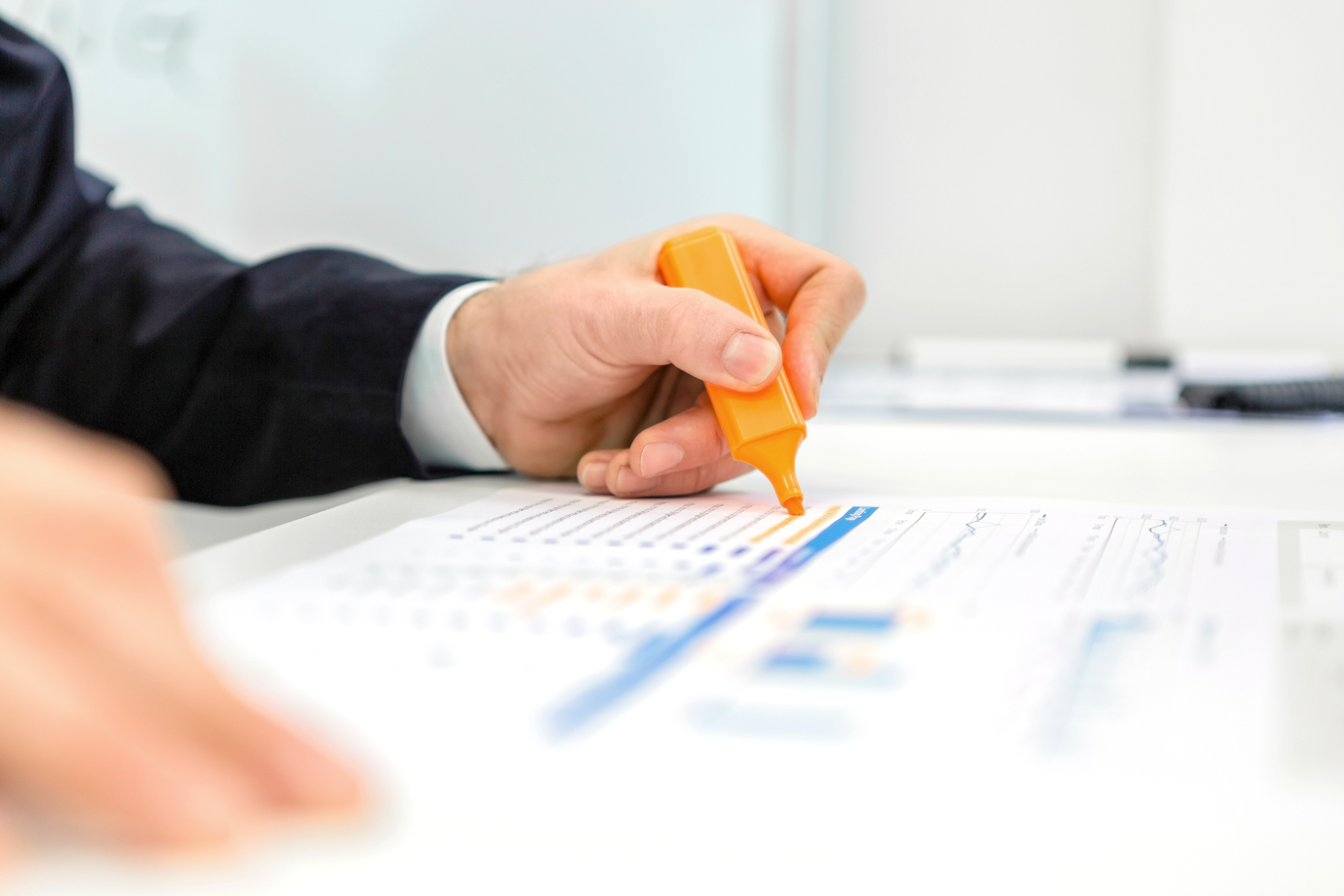Linke Hand eines Geschäftsmanns markiert mit Textmarker in orange eine Zeile in Tabelle