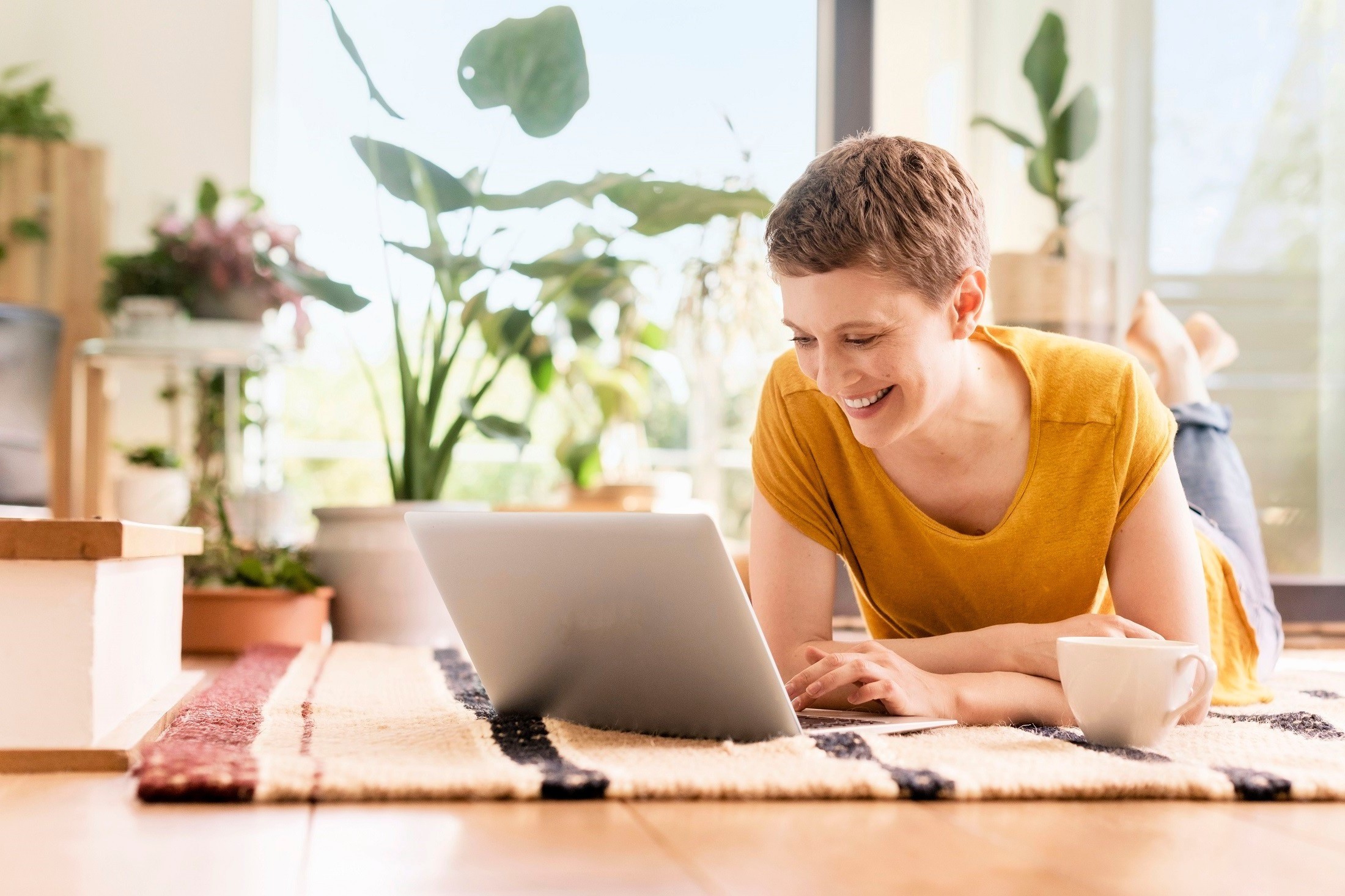 Frau liegt auf dem Boden mit einer Tasse und benutzt ihren Laptop