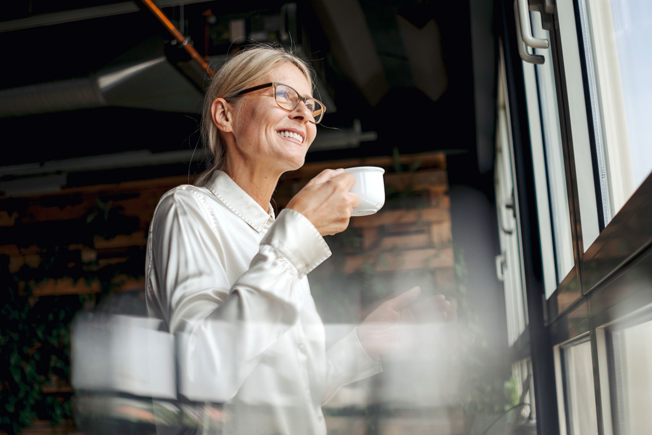 Lächelnde Geschäftsfrau schaut aus dem Fenster und hält eine Tasse Kaffee in der Hand