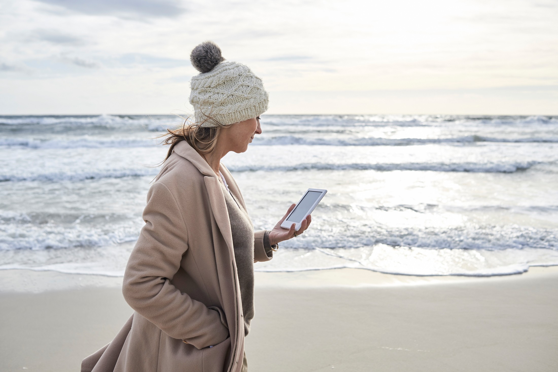 Frau mit Mütze läuft mit einem E-Book in der Hand am Strand entlang