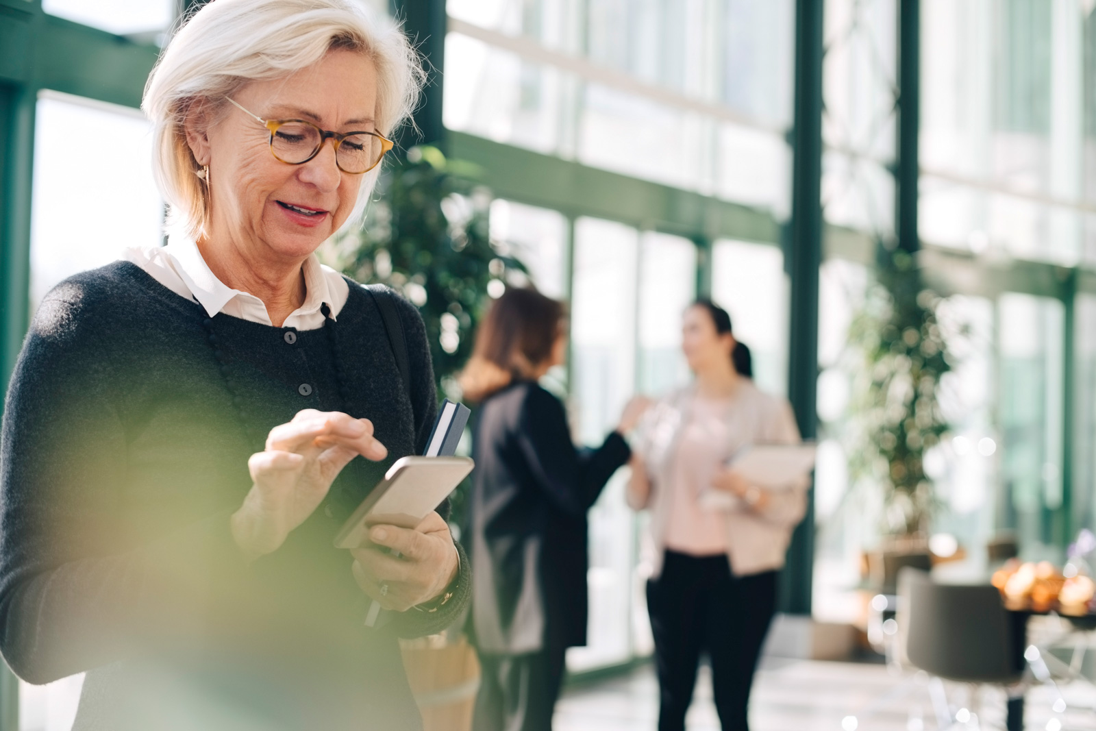 Senior Partnerin benutzt ihr Handy während Kollegen im Hintergrund diskutieren