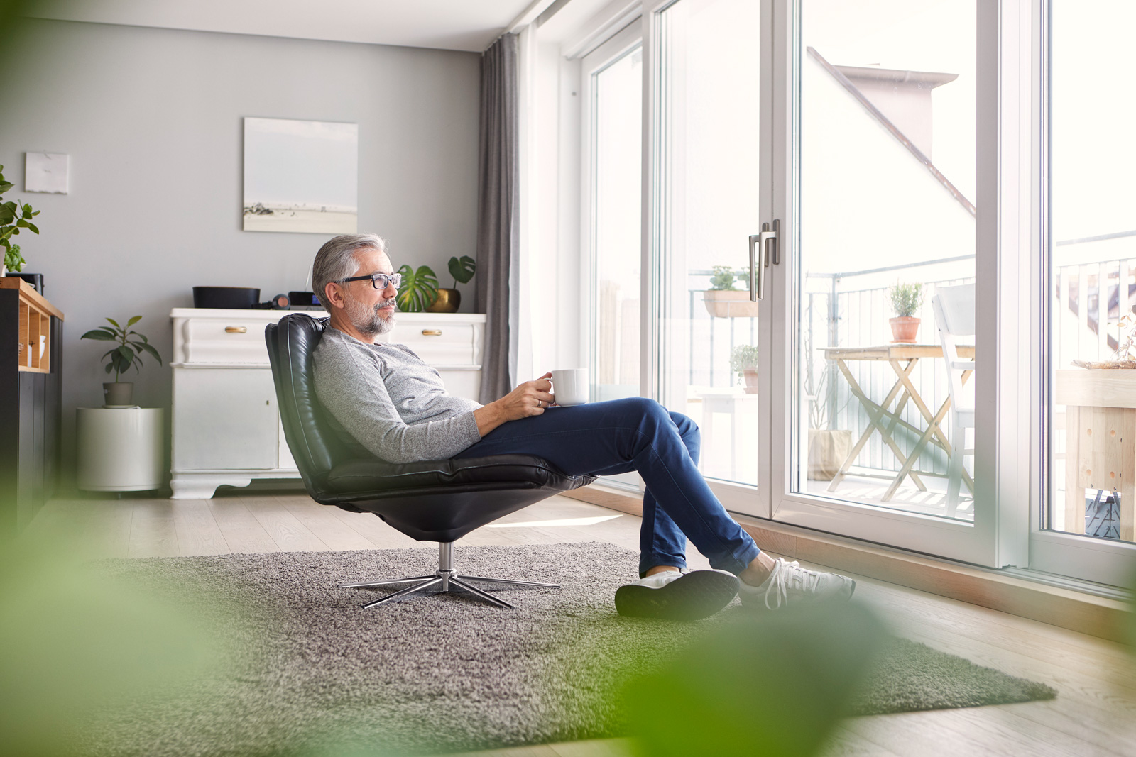 Älterer Mann sitzt in seinem Wohnzimmer in einem Lederstuhl mit einer Tasse Kaffee in der Hand